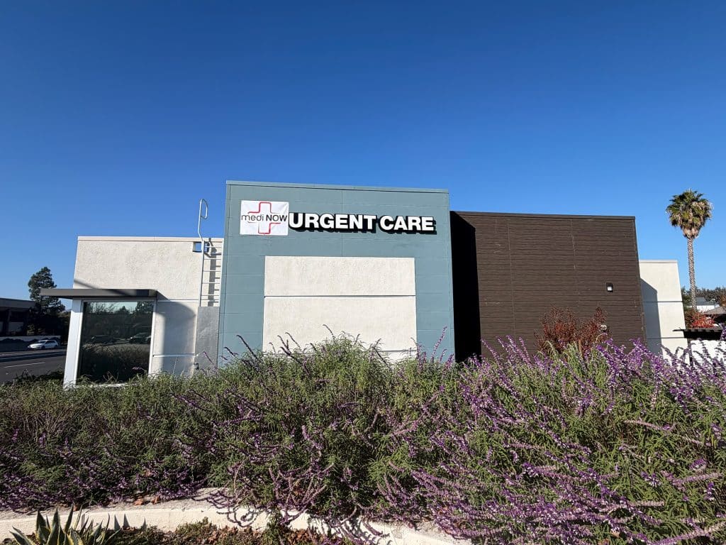 A modern urgent care clinic building with a gray and white exterior, a sign reading “medNOW URGENT CARE,” and purple flowering plants in front under a clear blue sky.