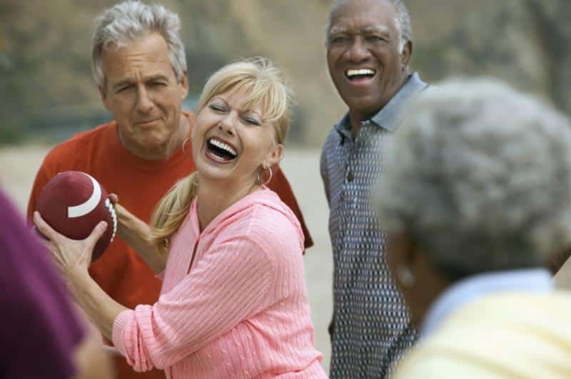 A group of older adults laugh and smile outdoors. One woman in a pink top holds a football, with three others around her, enjoying a playful moment together. The background is blurred, suggesting a park or natural setting.