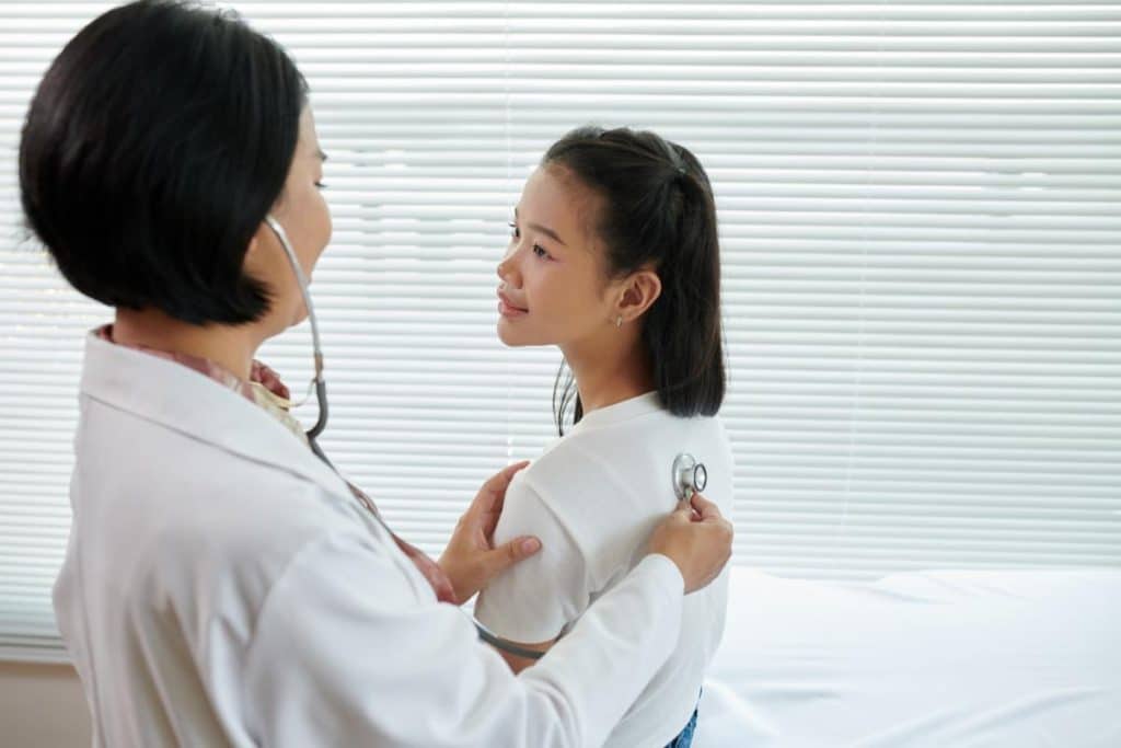 A doctor uses a stethoscope to listen to a young girl's back during a medical examination in a brightly lit room with window blinds. The girl sits on an exam table, looking calm and attentive.