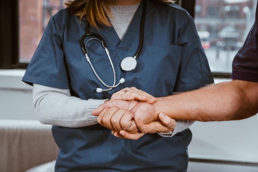 A healthcare worker in blue scrubs and a stethoscope gently holds a patient’s hand, offering comfort and support in a medical setting.