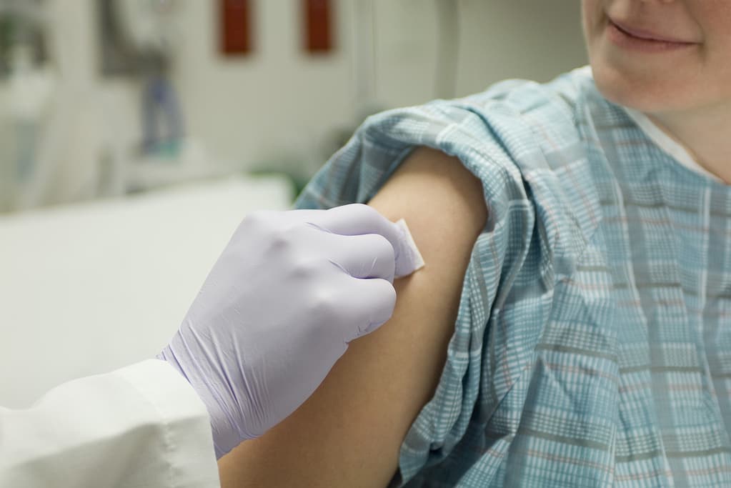 A healthcare worker wearing a purple glove cleans a patient’s upper arm with a cotton pad. The patient, dressed in a hospital gown, is smiling and sitting in a medical setting.