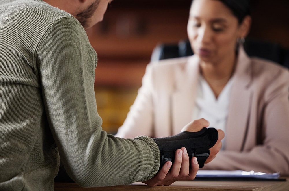 A person with a wrist brace sits across a desk from a professional woman in business attire, discussing paperwork in an office setting.