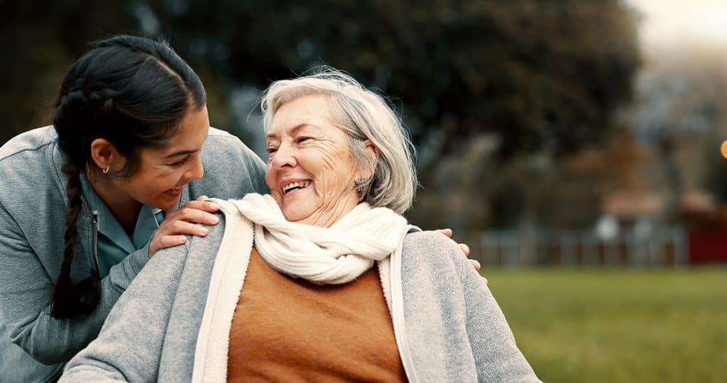A young woman smiles and leans toward an elderly woman in a scarf and sweater who is sitting in a wheelchair, both laughing together outdoors in a park.