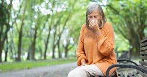 An older woman in an orange sweater sits on a park bench, holding a tissue to her nose, appearing to sneeze or have allergies, with green trees in the background.