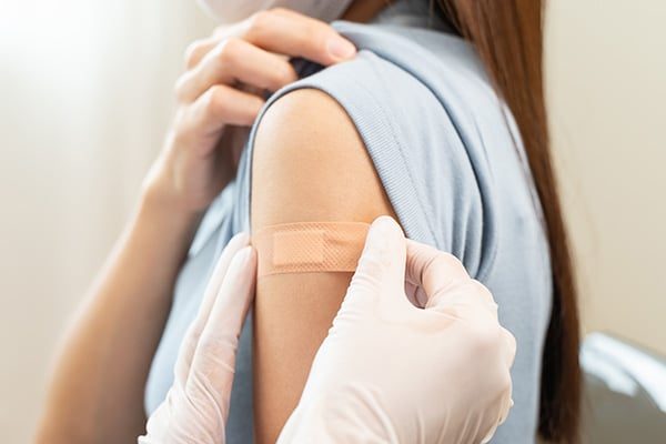 A person wearing a blue shirt has a bandage on their upper arm. Another person, wearing white gloves, is gently pressing the bandage, likely after giving a vaccination.
